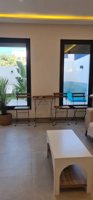 Un salon moderne avec vue sur la piscine, deux tables en bois, une table basse blanche et un canapé.