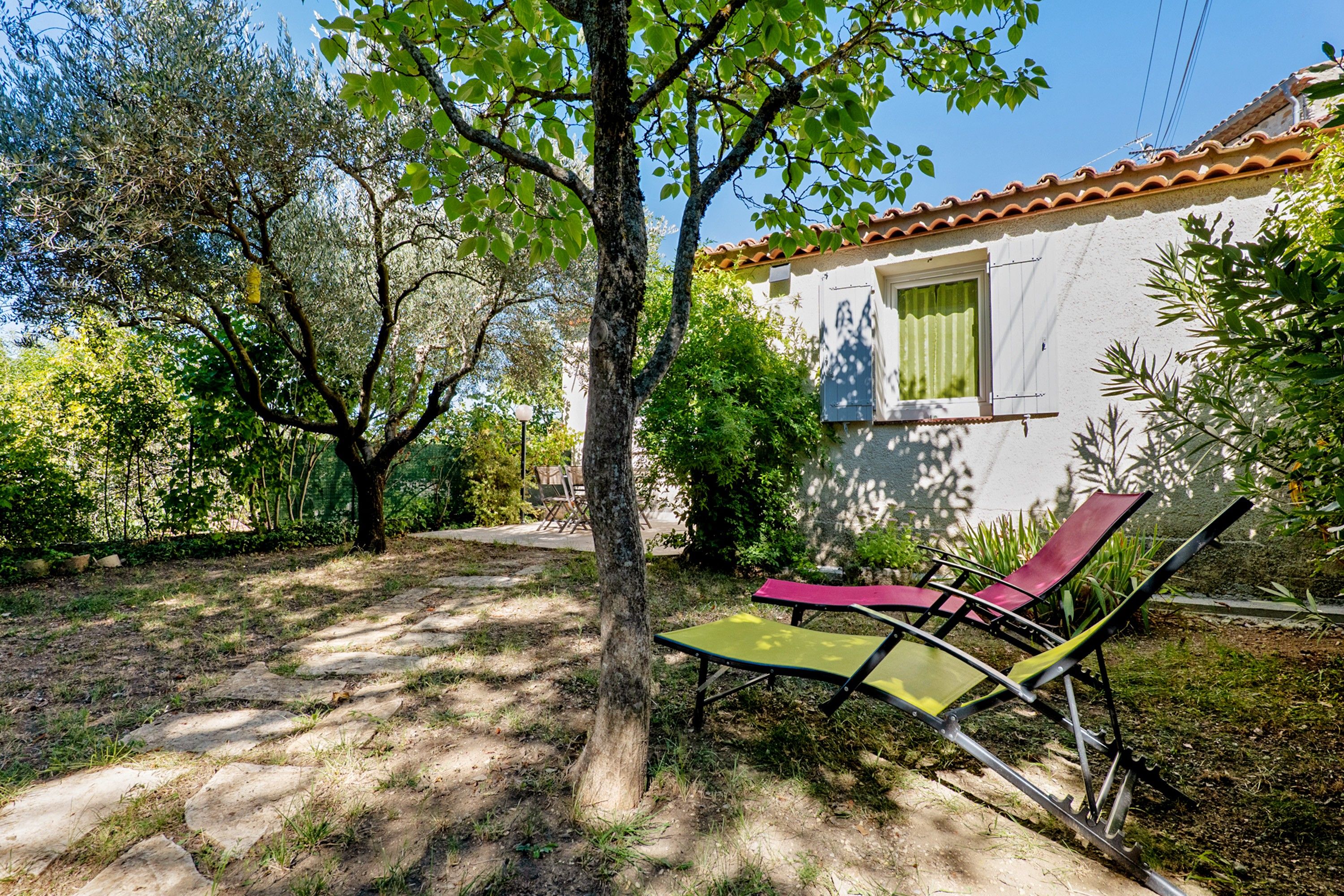 Una casa con fachada blanca, techo de tejas y un jardín con un árbol y dos tumbonas.