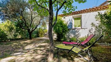 Una casa con fachada blanca, techo de tejas y un jardín con un árbol y dos tumbonas.