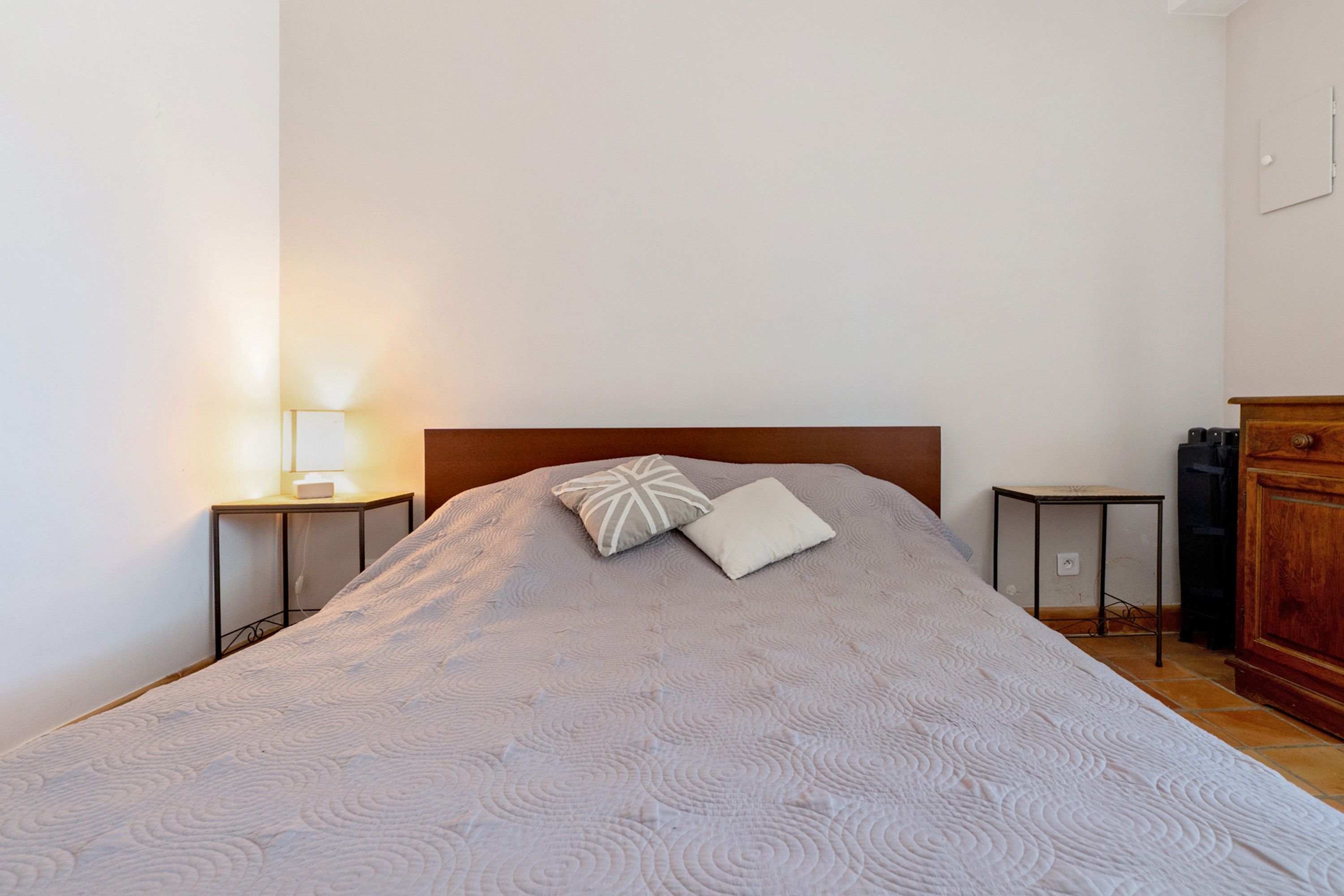 Ein ordentlich bezogenes Bett mit einem gemusterten Überwurf und zwei Kissen, ein Nachttischchen mit einer Lampe sowie eine Holzkommode in einem schlichten Zimmer.