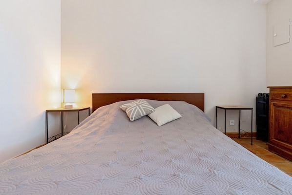 Un lit bien fait, avec une couvre-lit à motifs et deux oreillers, une table de chevet avec une lampe, et une commode en bois dans une pièce sobre.