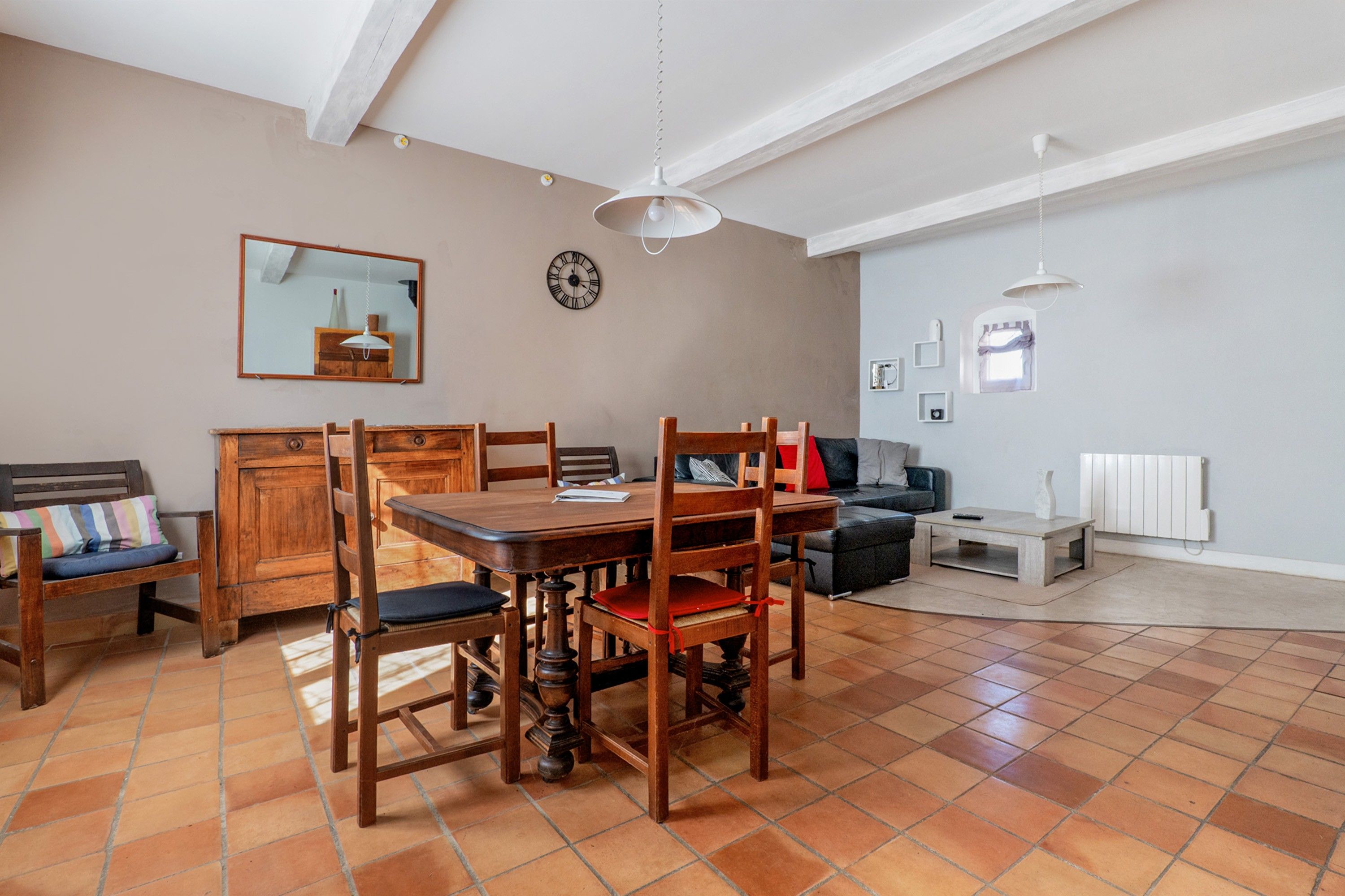 A dining area with a wooden table and chairs, a mirror, a clock, and a sofa in a living room with a tiled floor.