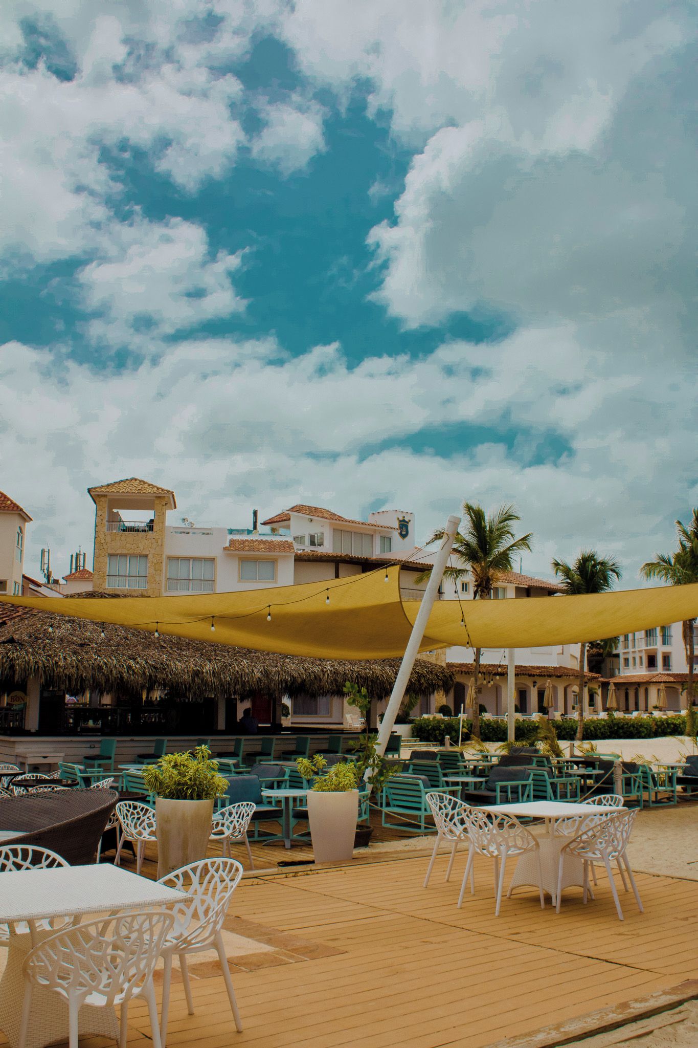 An outdoor seating area with tables and chairs, a thatched roof structure, and buildings in the background.