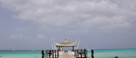 A wooden pier leading to a thatched-roof structure with white lounge chairs.