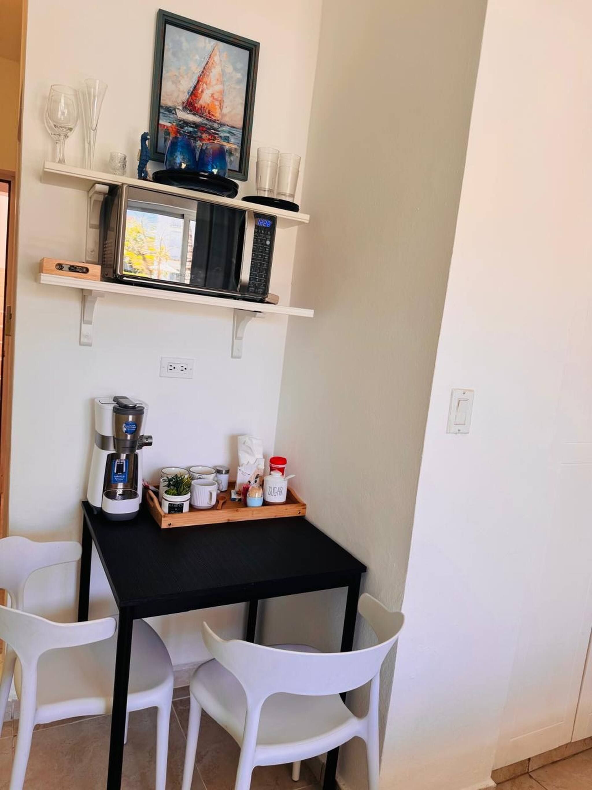 Eine kleine Küchenzeile mit Mikrowelle, Kaffeemaschine und einem Regal mit Gläsern und einem Gemälde. Zwei weiße Stühle und ein kleiner Tisch mit Küchenutensilien sind vorhanden.