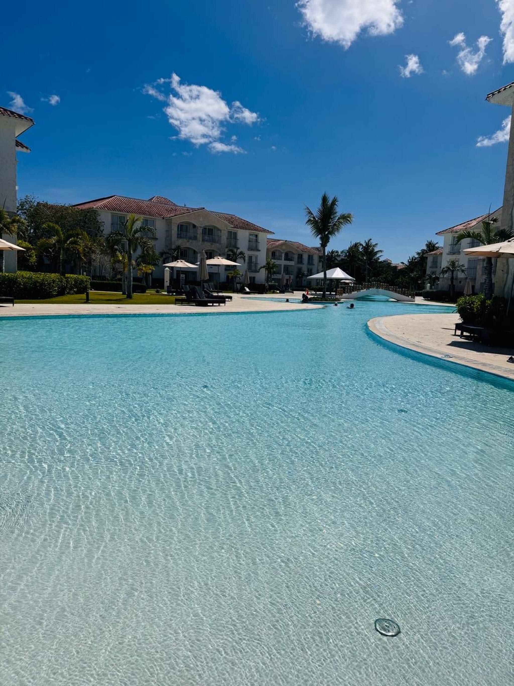 A clear blue pool with lounge chairs and umbrellas, surrounded by buildings with a tiled roof.