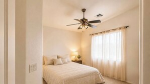 Ein Schlafzimmer mit einem Bett, einem Nachttisch, einer Lampe, einem Deckenventilator und einem Fenster mit Vorhängen.