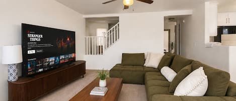 A living room with a flat-screen TV, a wooden coffee table, a green sofa, and a ceiling fan.