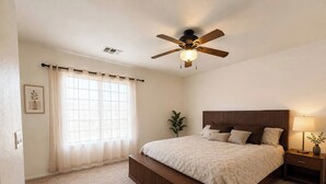 Ein Schlafzimmer mit einem Bett, einem Nachttisch, einer Lampe, einem Deckenventilator und einem Fenster mit Vorhängen.