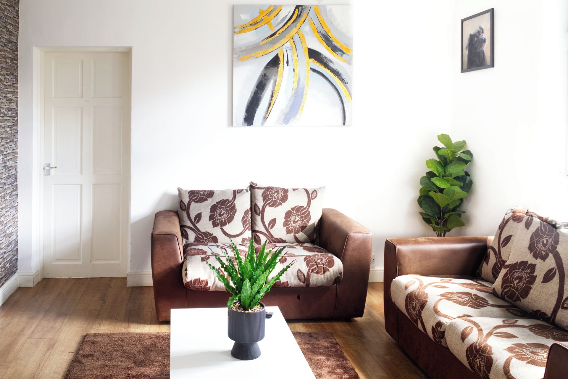 Ein Wohnzimmer mit einem braunen Ledersofa, einem weißen Couchtisch, einer Topfpflanze und einem Wandbild.
