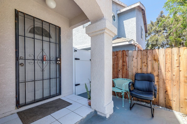 A gated entrance with a metal gate, a concrete pillar, a wooden fence, and a patio area with a chair and a small table.