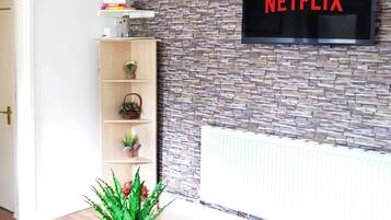 Een woonkamer met een muur met steenmotief, een wandgemonteerde televisie waarop 'NETFLIX' staat, een witte salontafel met een potplant en een boekenkast met decoratieve objecten.