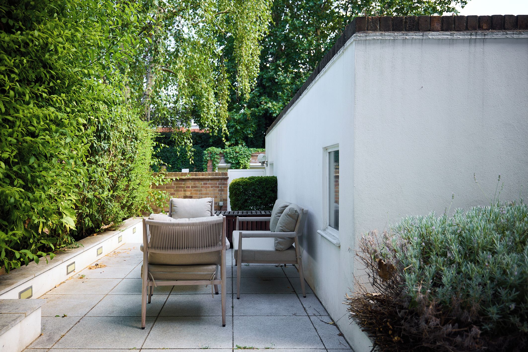 A patio with two chairs and a table, surrounded by greenery and a white building.