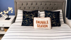 Ein Schlafzimmer mit einer gepolsterten Kopfteile, einem gerahmten Kunstwerk an der Wand und einem Kissen mit dem Aufdruck 'SNUGGLE SEASON'.