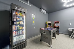 A vending machine with a 24-hour service sign, a foosball table, and two arcade games in a room with grey walls and a tiled floor. - Solterra Resort Home w/ AI Vending & Private Pool! (Davenport)