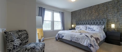 A bedroom with a large bed, a chair, a nightstand, and a window with curtains.