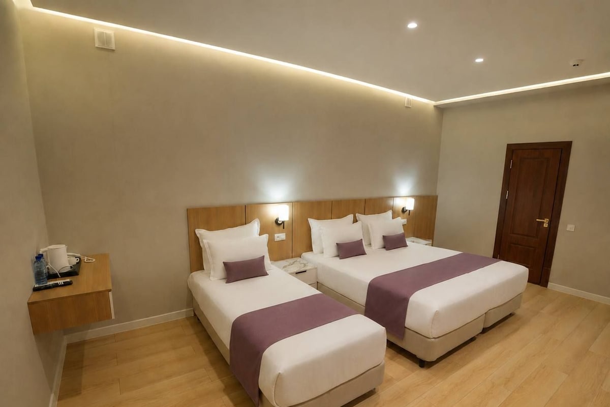 A hotel room with two beds, a wooden nightstand, and a door.