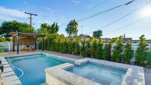 Ein rechteckiger Pool mit Holzterrasse und einer Pergola, umgeben von einem angelegten Garten und Wohngebäuden.