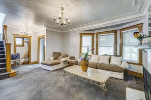A spacious living room with a high ceiling, a chandelier, a fireplace, and a large window with blinds.