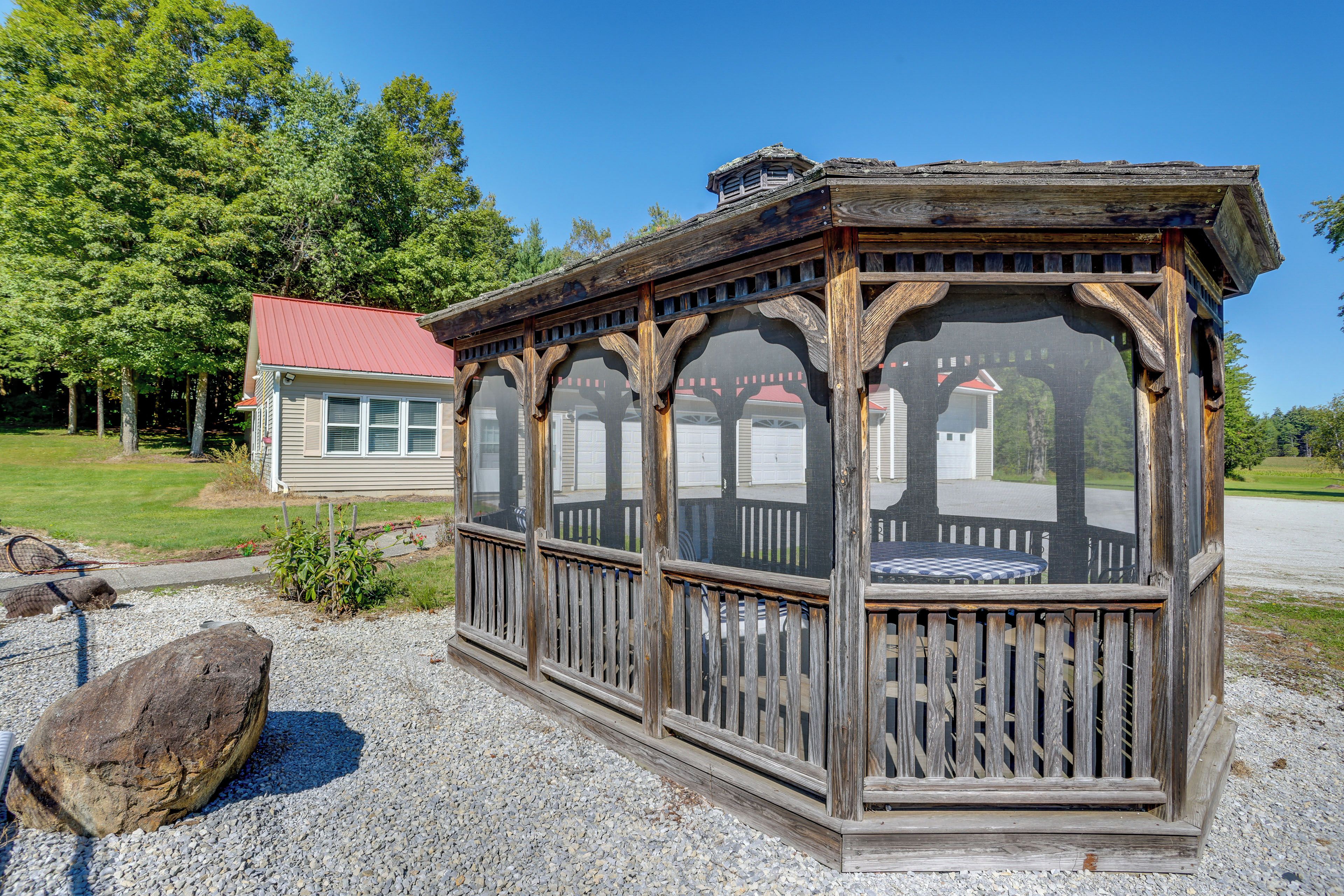 A wooden gazebo with a covered seating area, surrounded by gravel and a large rock.