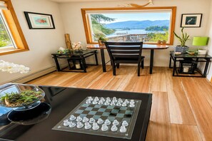 A room with a chessboard on a table, a desk with a chair, a window with a view of a lake and mountains, and a plant on the windowsill. - Ketchikan Oceanfront Retreat | Dock On-Site, Beach (Ketchikan)
