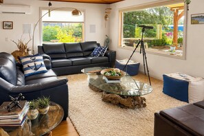 A living room with a black leather sofa, a glass coffee table, and a view of greenery outside. - Ketchikan Oceanfront Retreat | Dock On-Site, Beach (Ketchikan)