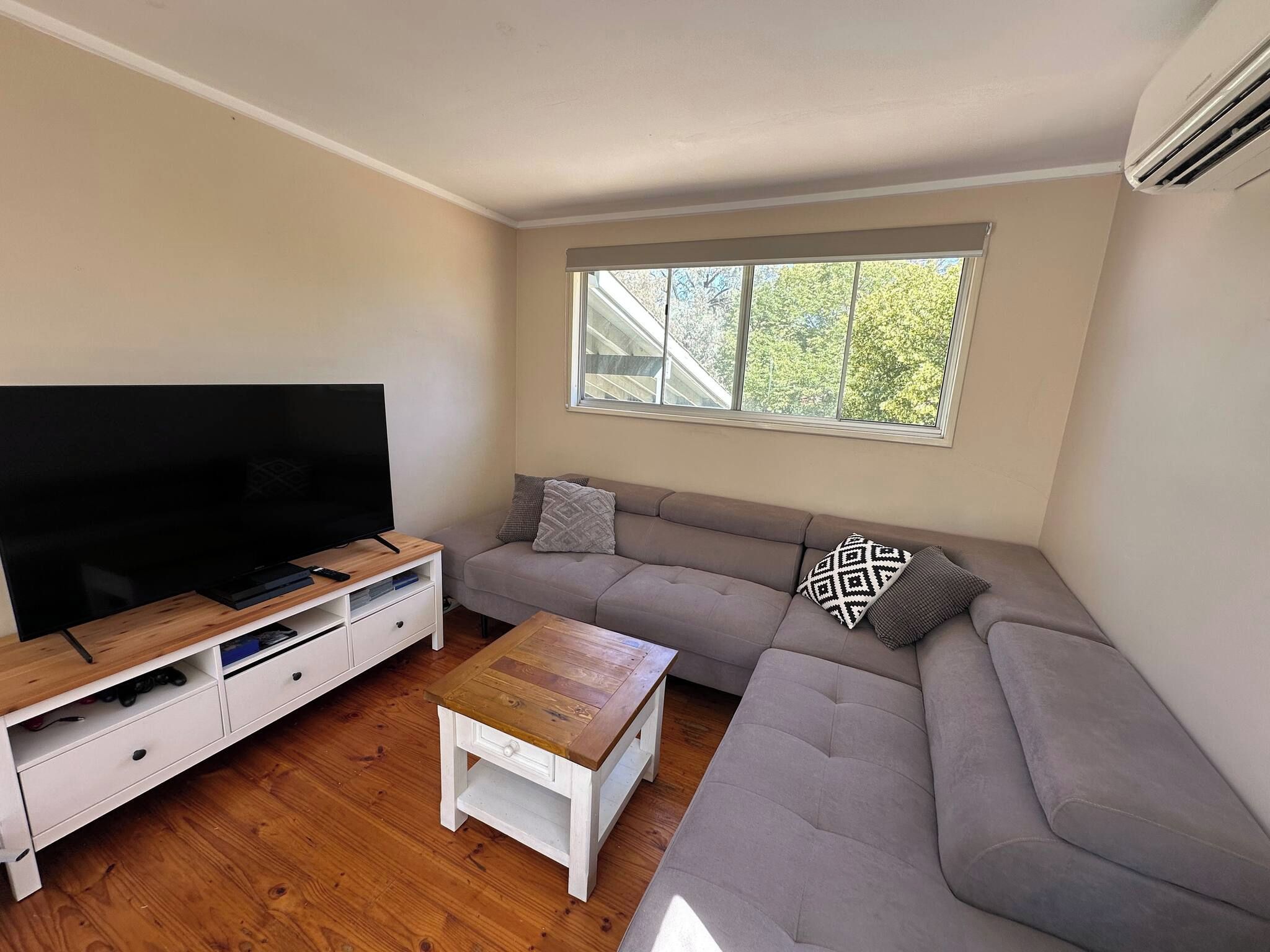 Een woonkamer met een televisie op een houten dressoir, een grijze bank met gekleurde kussens, een bijzettafeltje en een raam met uitzicht op bomen.