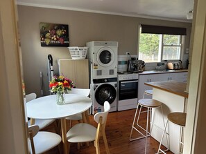 A compact kitchen with a washing machine, a coffee maker, and a dining area with a table and chairs. - Locum Welcome Home - Close to Canberra Hospital (Garran)