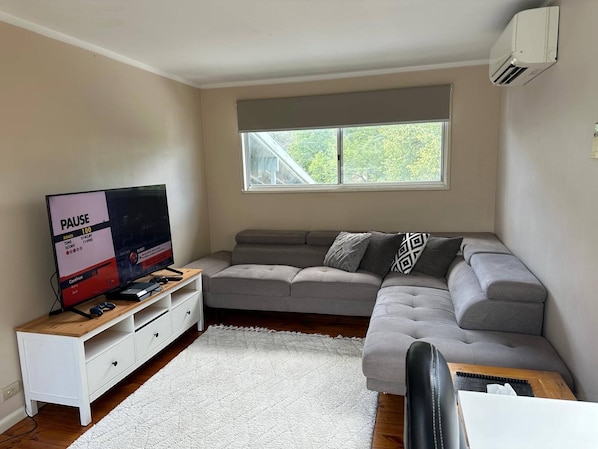 A modern living room with a grey sofa, a television on a wooden stand, a window with a view of trees, and an air conditioning unit on the wall. - Locum Welcome Home - Close to Canberra Hospital (Garran)