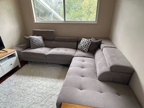 A modern living room with a sectional sofa, a small white dresser, and a window with a view of trees. - Locum Welcome Home - Close to Canberra Hospital (Garran)