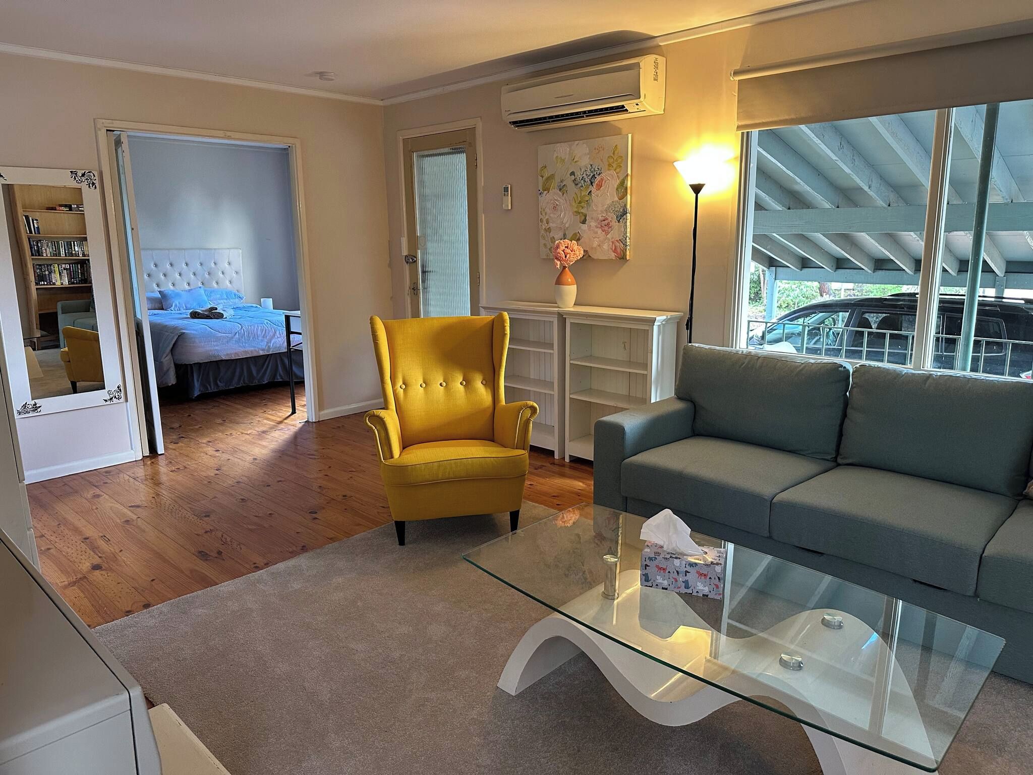 Ein Wohnzimmer mit einer Couch, einem gelben Sessel, einem Glastisch und einem Bett im angrenzenden Zimmer.
