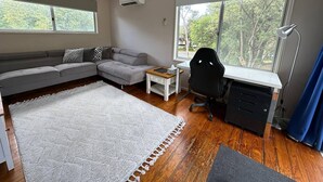 Una sala de estar moderna con un sofá seccional, una mesita, un escritorio con una silla, un ventanal grande y un equipo de aire acondicionado.