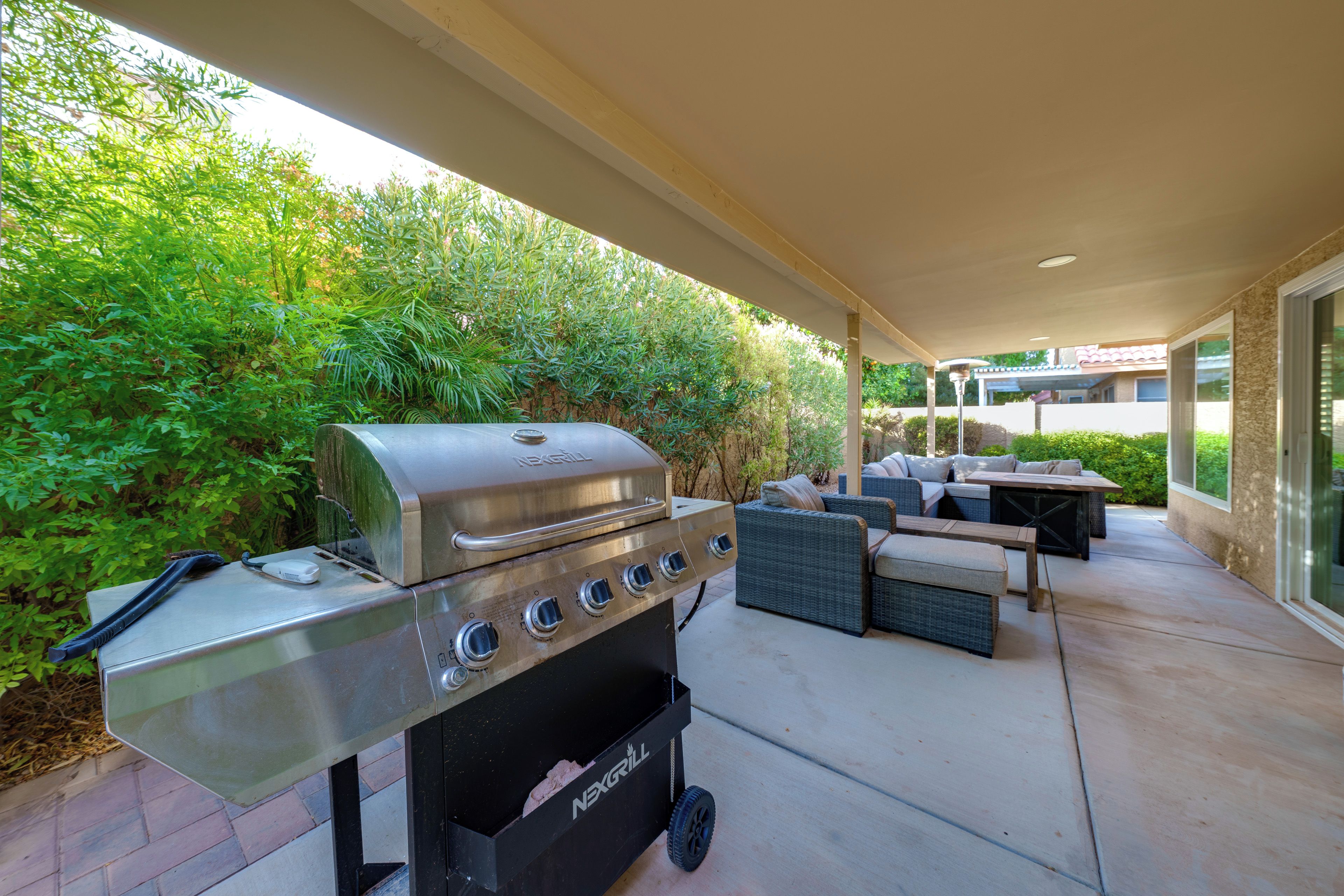 An outdoor patio with a stainless steel grill, wicker furniture, and a covered area with a view of greenery.