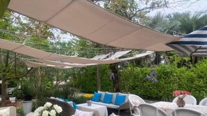 Eine Außenpatio-Terrasse mit Tischen und Stühlen, die von einer Markise beschattet und von viel Grün umgeben ist.