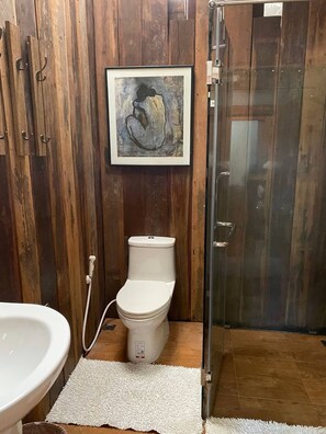 A bathroom with a wooden wall, a framed abstract painting, a white toilet, a glass shower enclosure, and a white sink. - the NOW by Kru Se Cottage (Chiang Mai)