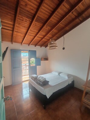 A bedroom with a wooden ceiling, a bed with white linens, a hanging light fixture, and a view through an open door.