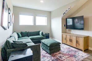 A living room with a sectional sofa, ottoman, wooden cabinet, flat-screen TV, and two windows with blinds. - Family Getaway, 5 Mi to Silver Dollar City (Branson)