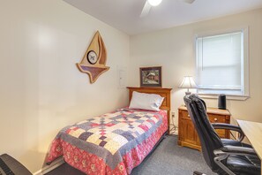Een slaapkamer met een bed, een houten hoofdbord, een sprei, een klok, een foto, een bureau, een stoel en een raam met jaloezieën.