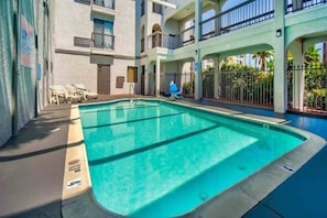 Una piscina con superficie de agua azul cristalina, rodeada por un edificio de varios niveles con balcones y un paseo techado.