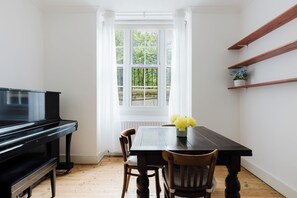 Ein Zimmer mit einem Klavier, einem Esstisch mit Stühlen, einem Fenster mit Vorhängen und Holzregalen.