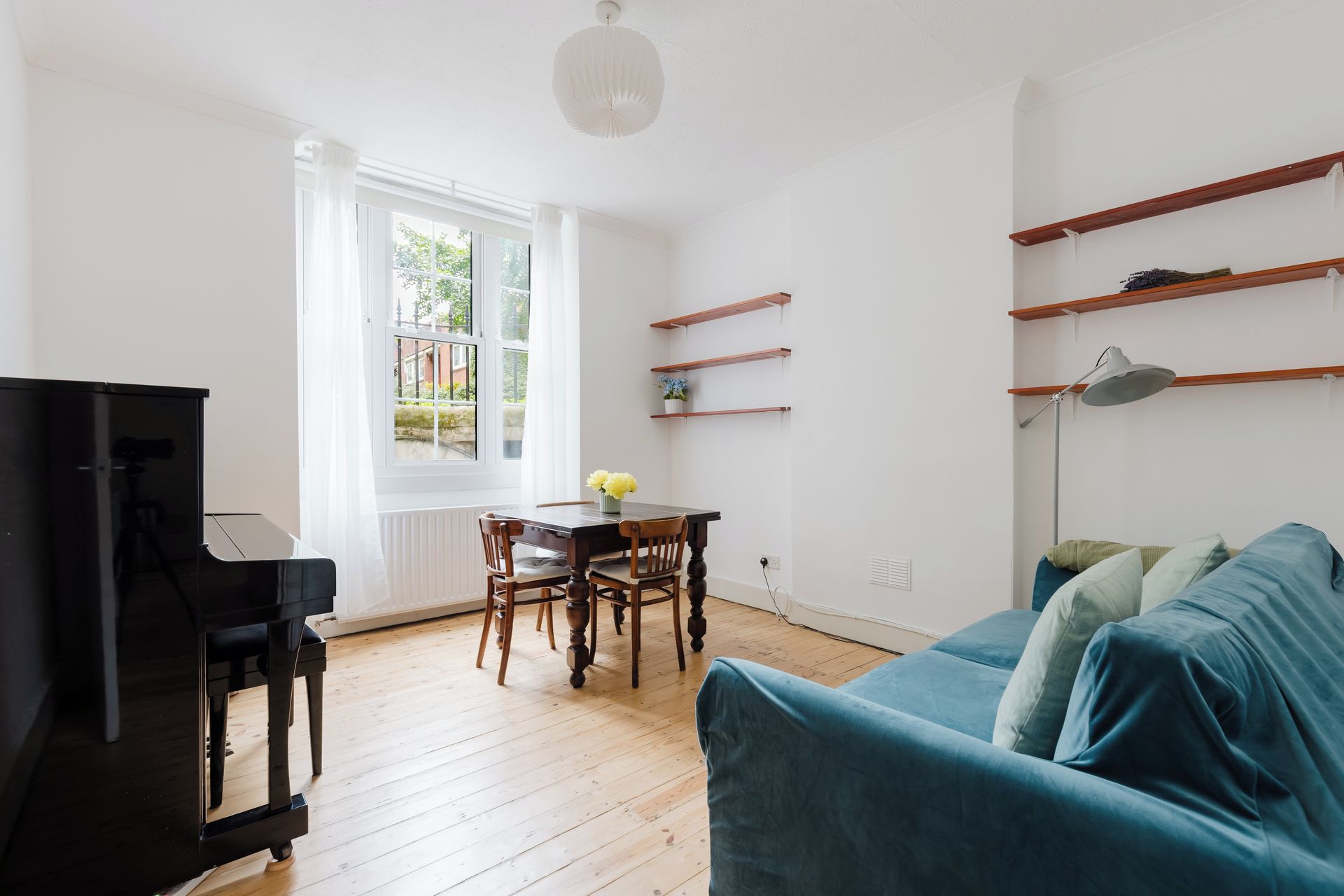 A living room with a piano, a blue sofa, wooden flooring, and a window with a view of greenery.