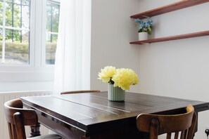 A dining table with a vase of yellow flowers, wooden chairs, a window with blinds, and a white wall with a shelf. - Timeless Retreat 5 Minutes from Tower Bridge (Greater London)