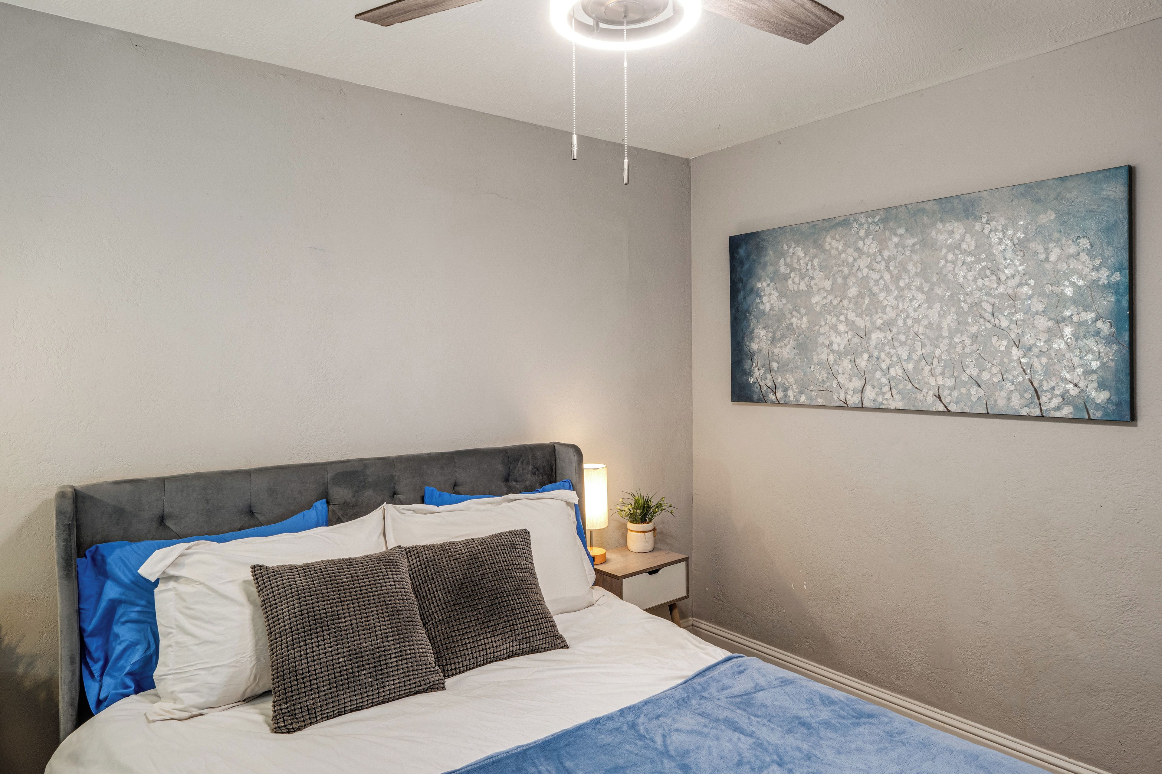 Un dormitorio con una cama, una mesita de noche, un ventilador de techo y un cuadro en la pared.