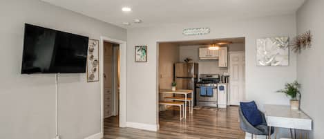A modern living room with a flat-screen TV, a dining area with a table and chairs, and a kitchen with stainless steel appliances.