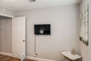 A room with a white door, a mounted flat-screen TV, a small white table with a gold wire shelf, and a painting on the wall. - Bakersfield Home w/ Workspace, Near Hospitals (Bakersfield)