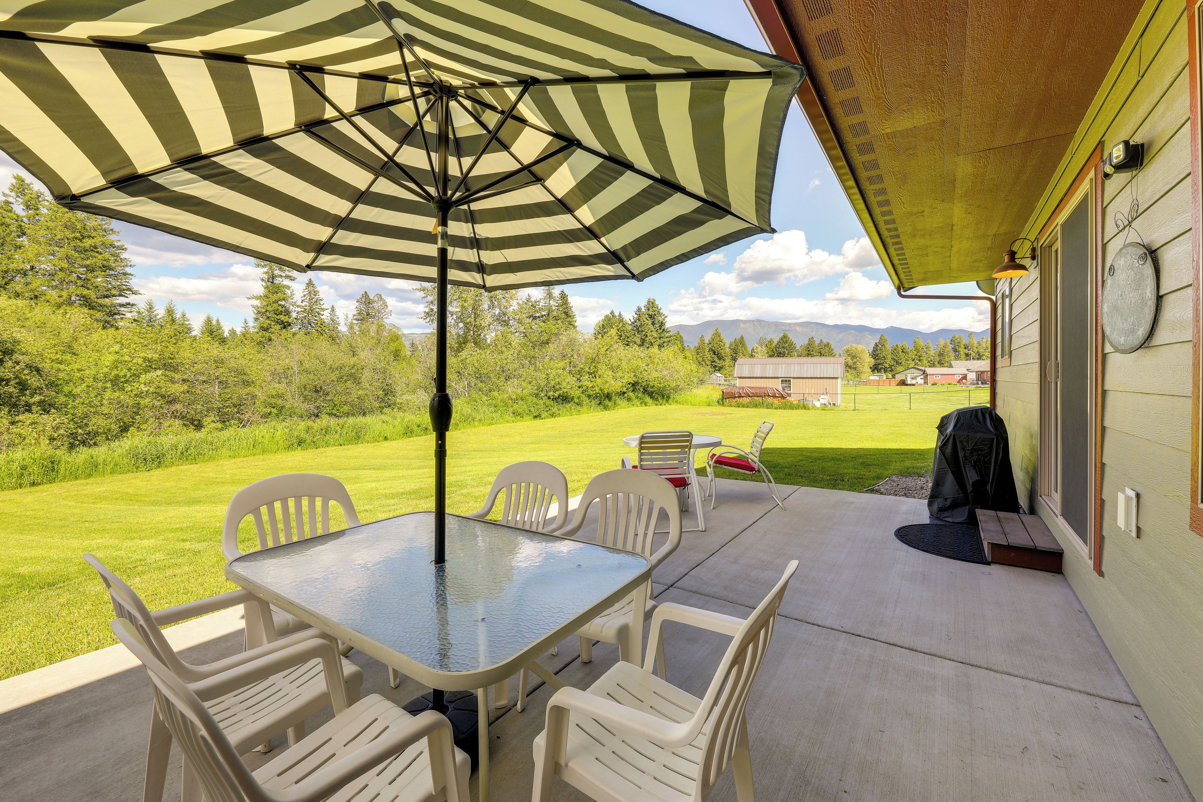 An outdoor patio with a table and chairs, a striped umbrella, and a view of a grassy yard and mountains.