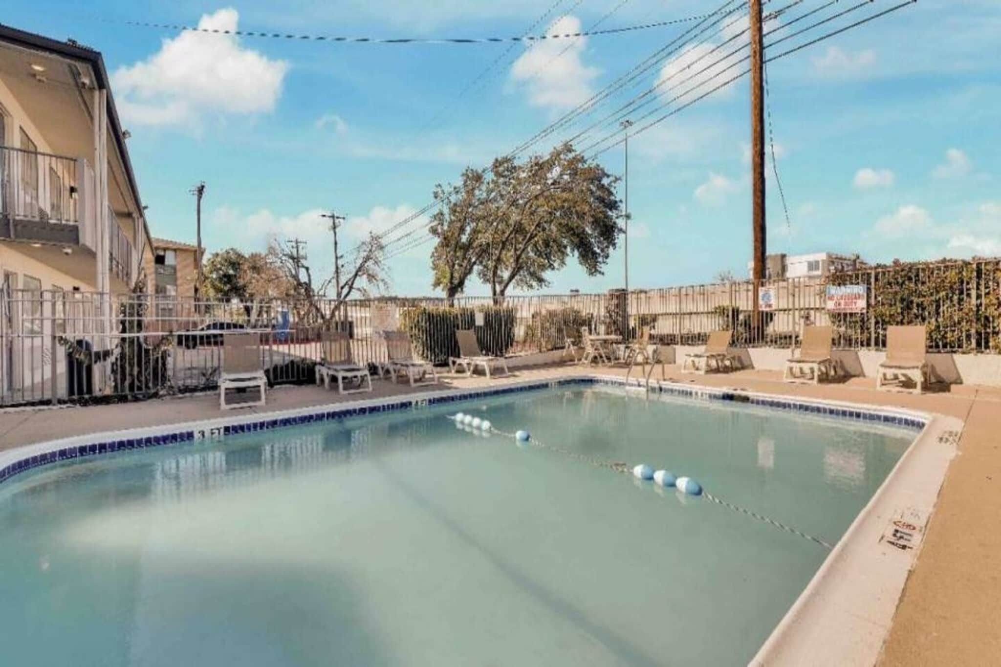 A swimming pool with lounge chairs and a fenced area, surrounded by buildings and trees.