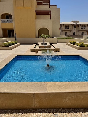 Ein rechteckiger Pool mit einer Fontäne in der Mitte, umgeben von einer gepflasterten Fläche mit Pflanzgefäßen und einem Gebäude mit Rundbogenfenstern im Hintergrund.