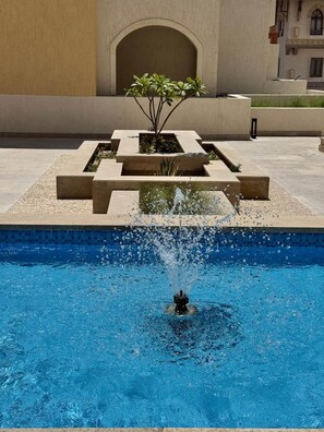 Ein Schwimmbecken mit Springbrunnen, umgeben von einem geometrisch gestalteten Pflanzgefäß mit Pflanzen.