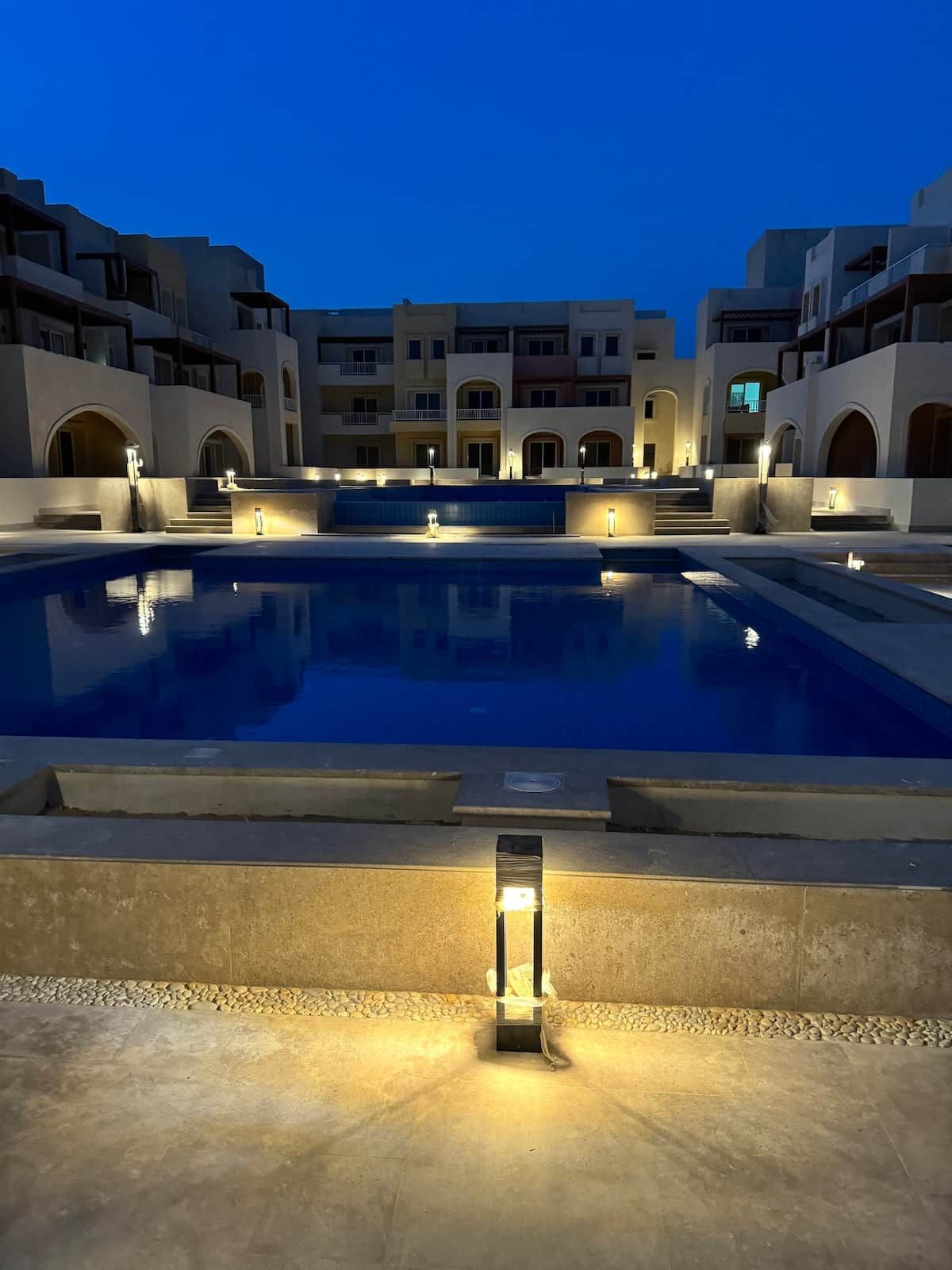 A swimming pool with a stone border and steps leading up to it, surrounded by buildings with balconies and arched doorways.